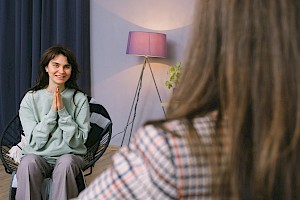 Kobieta siedzi w fotelu na przeciwko psychoterapeutki, w tle zasłonięte okno i lampa